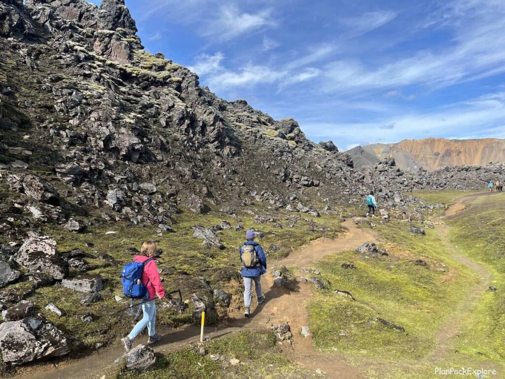 3 people hiking on the path along a lava field. Mountains of Landmannalaugar visible in the distance.