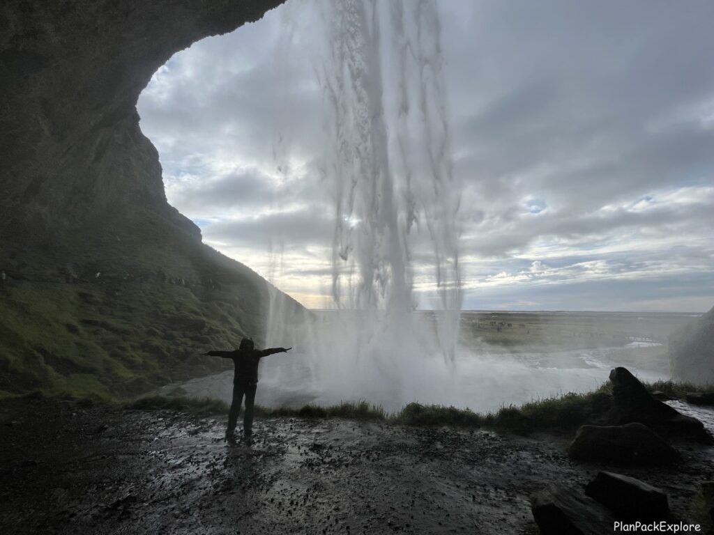 Article author standing with her arms stretched out to her sudes in a cavern inside Seljalandsfoss waterfall in Iceland. Waterfall visible behind her.