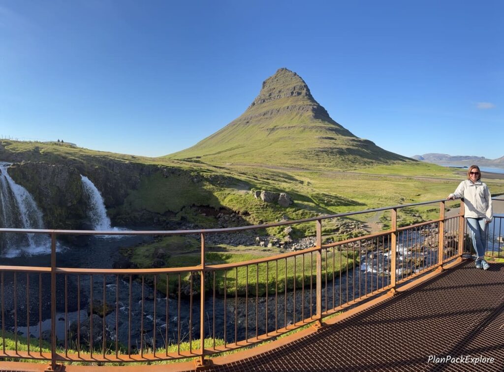 A pointy green mountain called Kirkjufell Mountain and a person standing on a viewing platform next to it on Snaefellsnes Peninsula in Iceland.