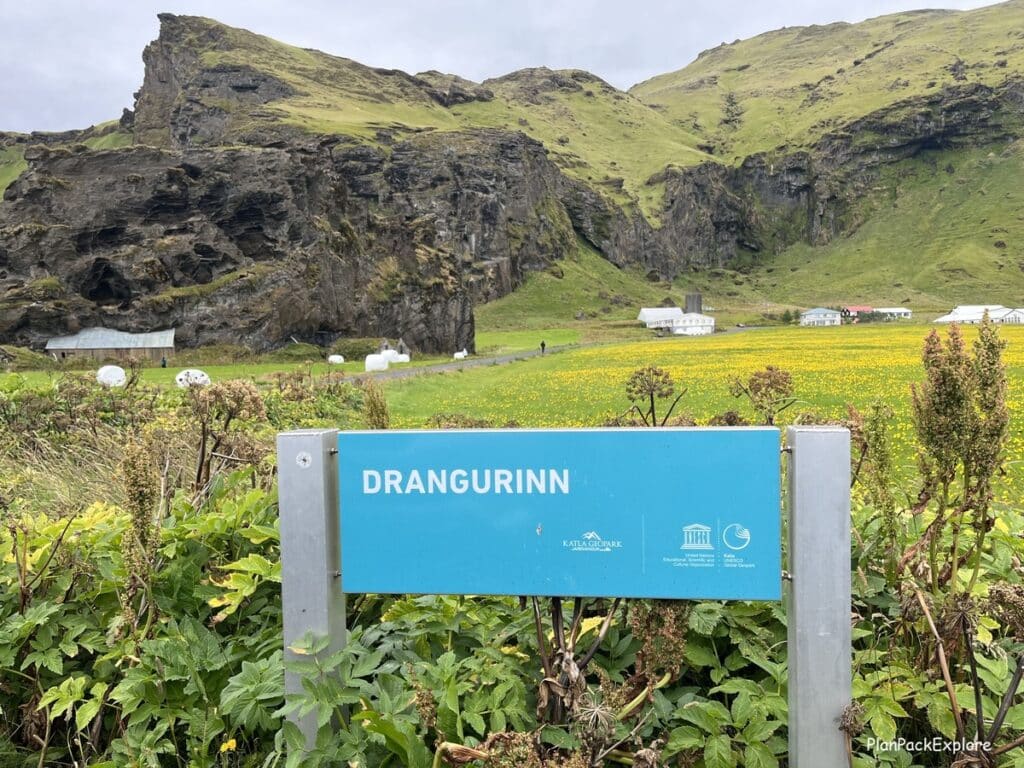 A blue sign with white letters for Drangurinn. Moss-covered mountains and Drangshlíð turf houses in the background.