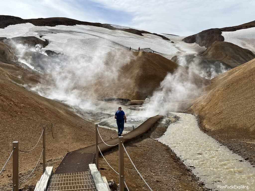 A person crossing a bridge over a steaming stream in Hveradalir Geothermal valley, near Kerlingarfjoll, Iceland. Steaming vents in the background.