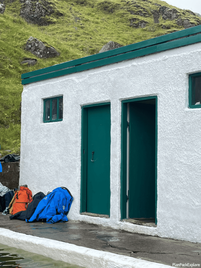 Changing room of Seljavallalaug swimming pool in Iceland - a small white building with green roof and doors and some backpackcs in front of it.