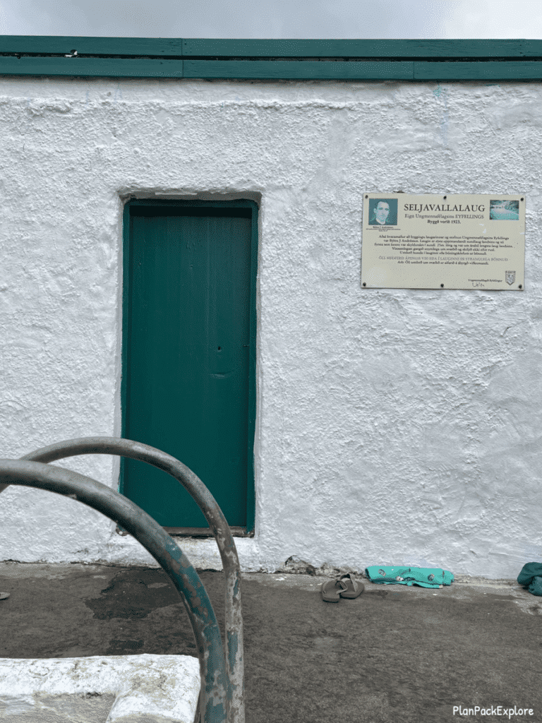 Changing room of Seljavallalaug swimming pool in Iceland - a small white building with green doors. A plaque with pool's history and founder info on the wall.