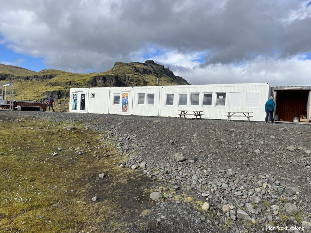 A small white building (Icelandic Mountain Guides base) on a gravel lot of a parking area for Solheimajokull glacier in Iceland.