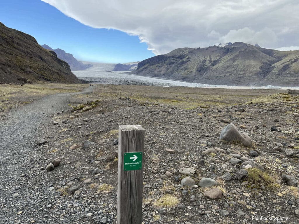 A gravel path leading to Skaftafel glacier in Iceland. A glacier and a trail marker post with a green sign on it are also visible.