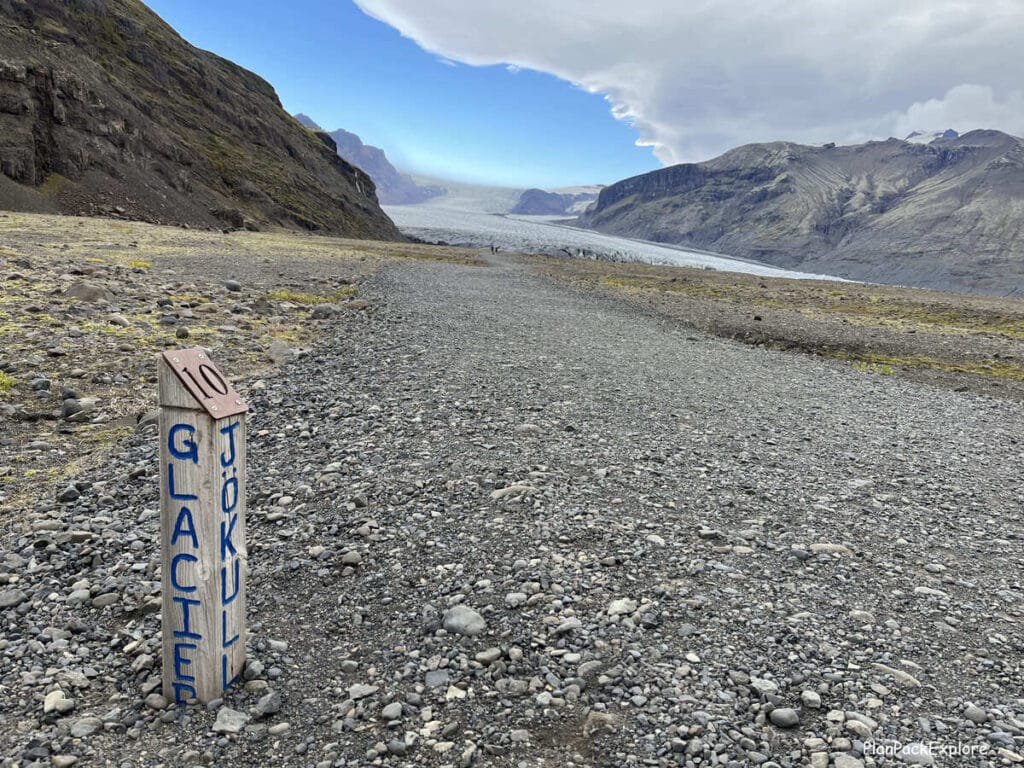 A gravel path leading to Skaftafel glacier in Iceland. A glacier and a numbered trail marker post with the words "glacier" and "jokull" on it are also visible.