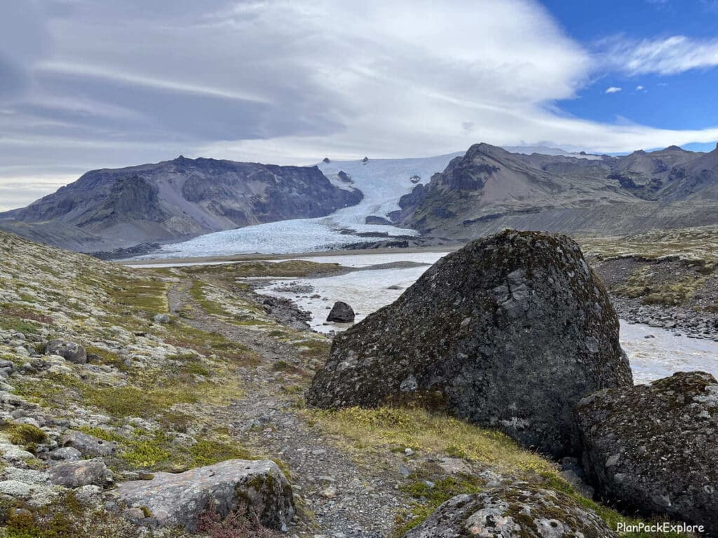 Kviarjokull glacier in Iceland - a view of a gravel path with a large boulder on the right side and a glacier with a small lagoon in front of it.