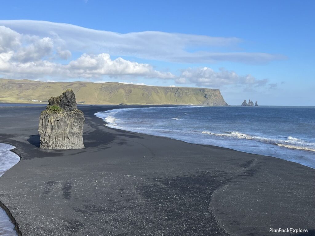 A tall pointy basalt rock formation on a black beach and a view od reynisdrangar sea stacks in the distance from dyrholaey in Iceland.