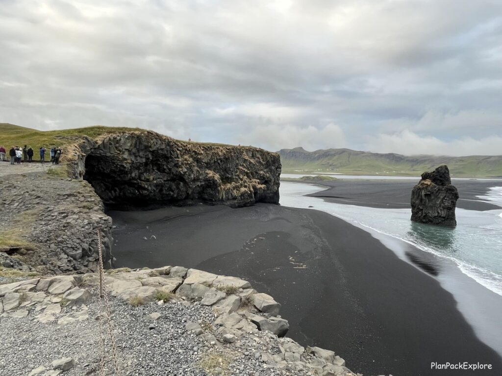 People watching puffins at Dyrholaey Cliffs in Iceland. A single basalt rock formation is standing in the ocean, near a black sand beach.