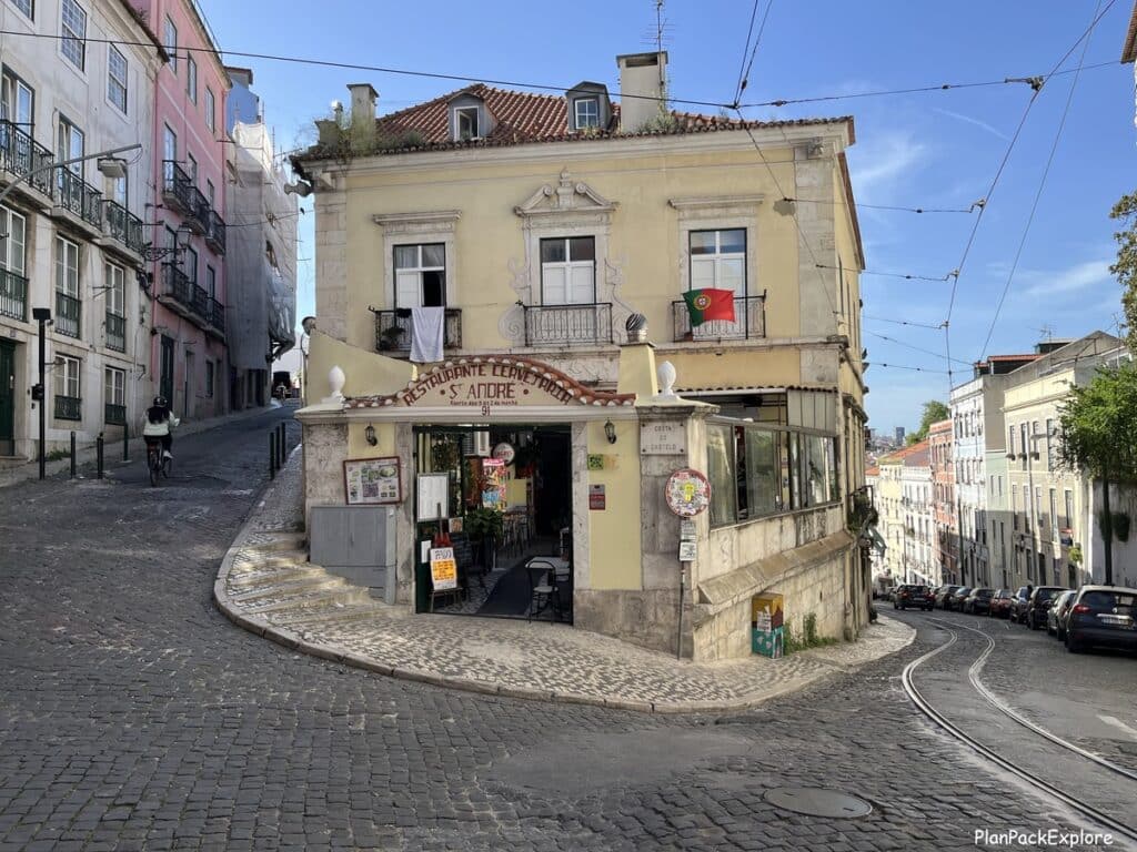 corber of a winding cobblestoned street in Lisbon, Portugal, with a cream-colored building in the middle.