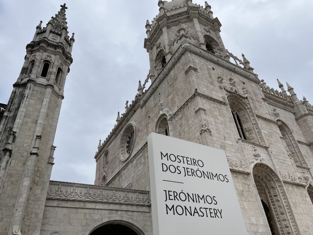A top part of a stone historic building with a sign saying Jeronimos Monastery in Lisbon.