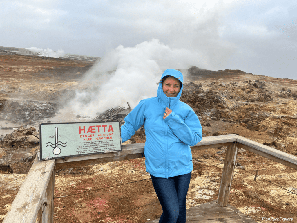 A person in a blue jacket standing next to a warning sign with a steaming hot spring in the back on reykjanes peninsula in iceland.