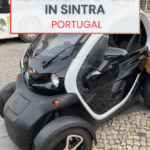 A black and white electric vehicle is parked on a cobblestone street in Sintra, Portugal, with text overlay promoting a guide to renting an e-car.