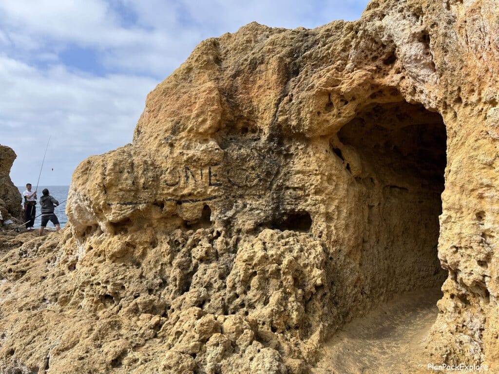 An entrance to a cave in Algar seco in the Algarve, Portugal, marked by a worn sign in black letters, spelling "A Boneca".