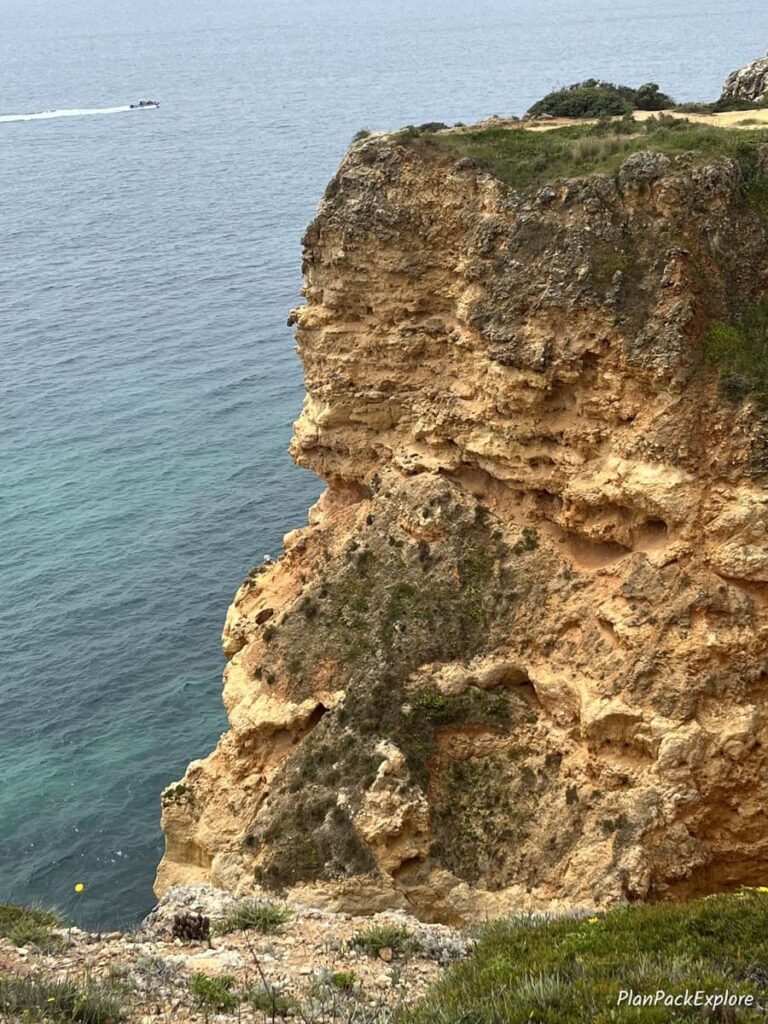 A tall cliff by the ocean with a small boat in the background, on the trail of the headlands in the Algarve, Portugal.