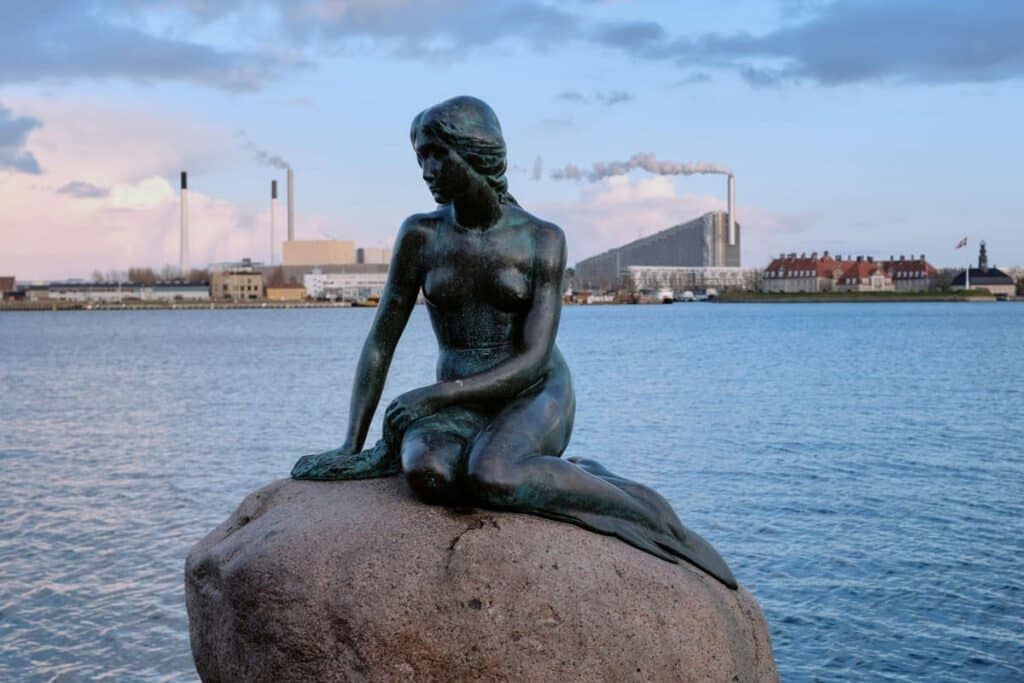 A statue of a little mermaid sitting on a large rock in the water in copenhagen.