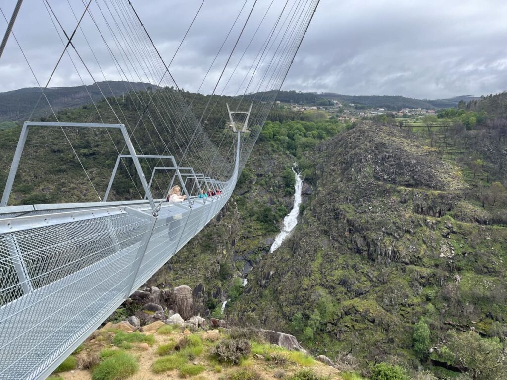 A person standing on the steel suspension bridge called 516 Arouca Bridge in Portugal, with a waterfall visible.