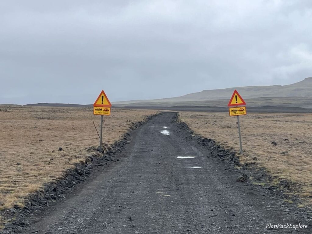 A gravel road with two yellow warning signs in Thorsardalur Valley, Iceland.
