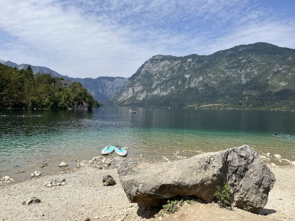 A blue lake surrounded by maountains with a large rock and two paddleboards on a small sandy beach by Lake Bohinj, Slovenia.