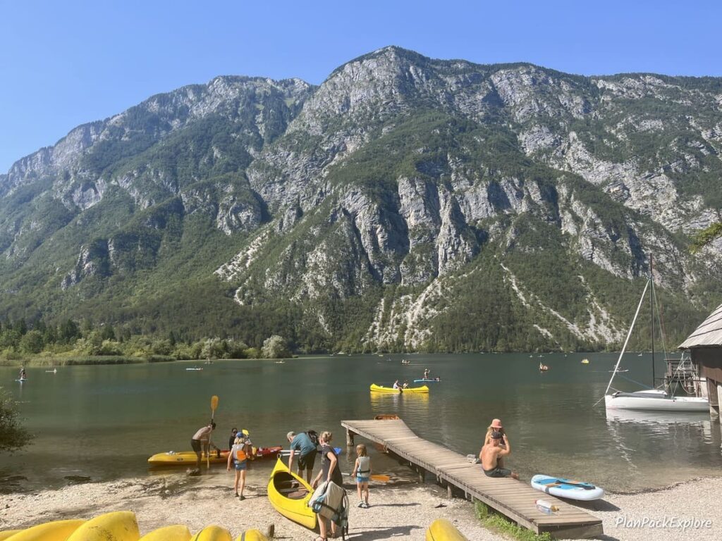 A small sandy beach with kayaks on the shore of Lake Bohinj in Slovenia, surrounded by mountains.