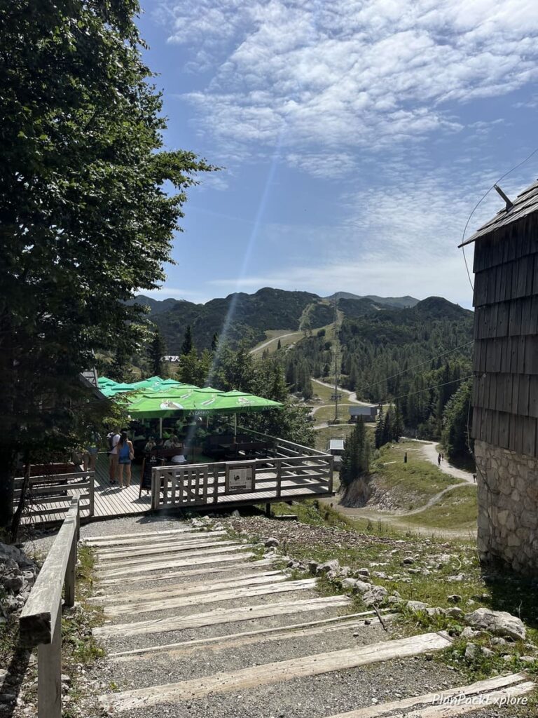 Wooden stairs leading to an outside sitting area with tables and green umbrellas of a restaurant at the top of Vogel cable car station in Slovenia.