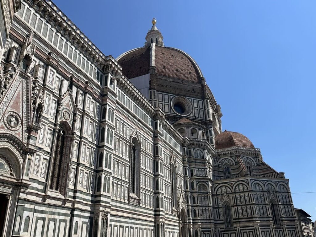 The image shows the exterior of the Florence Catherdal, featuring intricate marble details, with a crowd of people walking in the foreground during twilight.