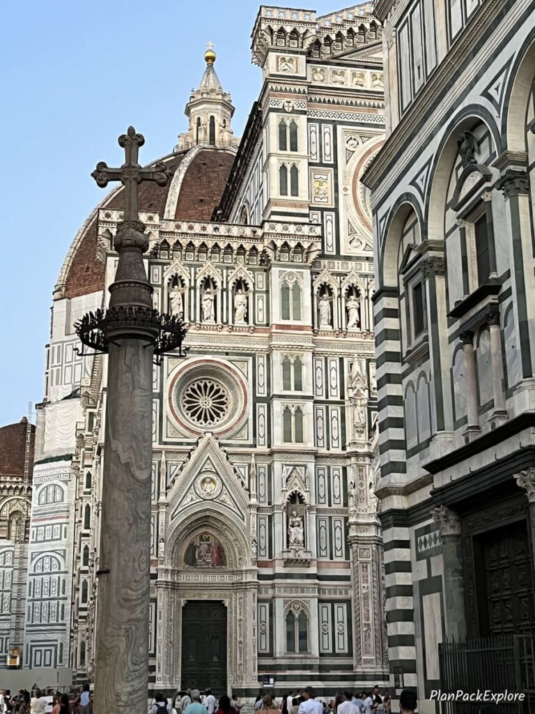 A partial view of the Florence Cathedral dome and a tall pillar with a bronze cross on top.