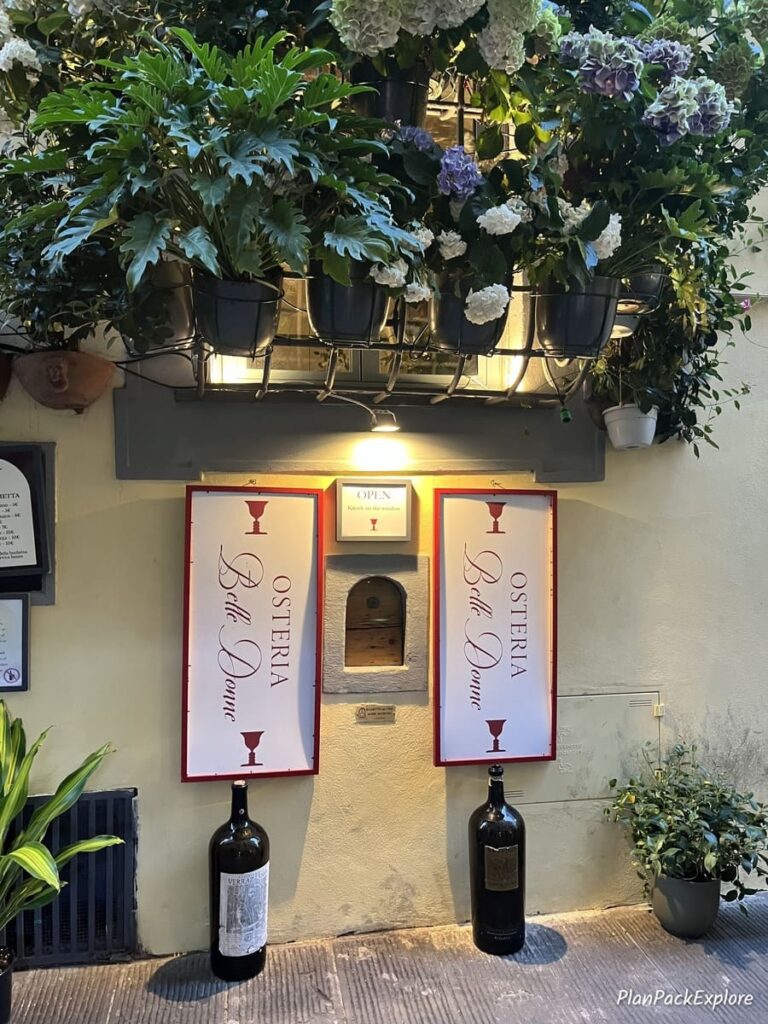 A wine window in Florence - a small opening in the wall with vines on top and white signs on both sides.