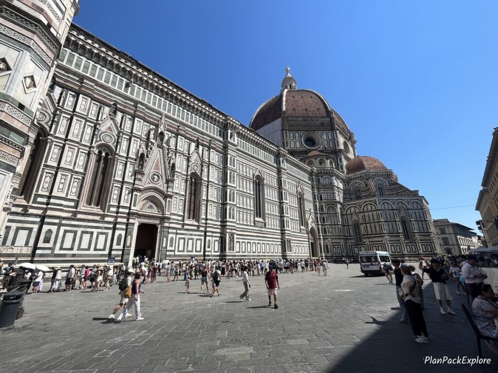The image shows the exterior of the Florence Catherdal, featuring intricate marble details, with a crowd of people walking in the foreground during twilight.