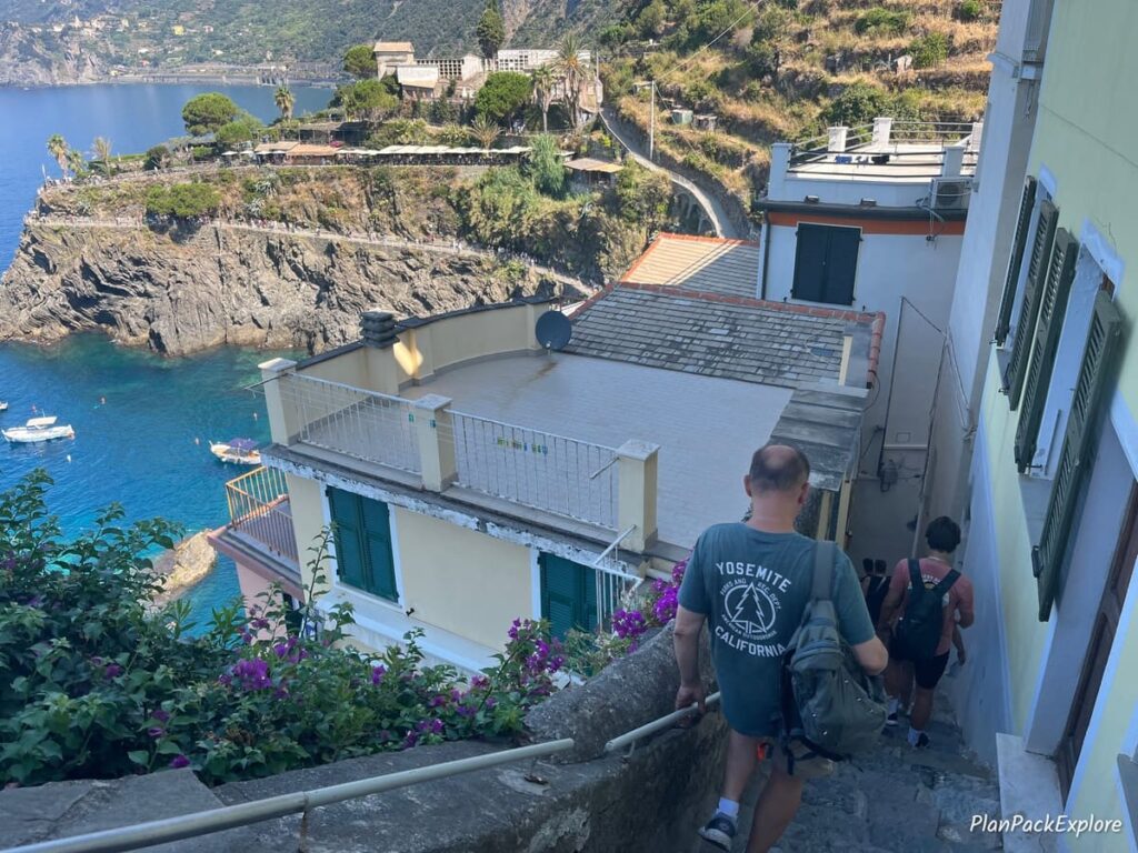 A person going down steep narrow stairs near a viewpoint, looking over the Ligurian Sea and houses and vineyards of Cinque Terre, Italy.