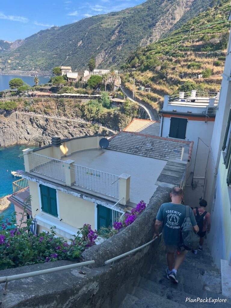 A person going down steep narrow stairs near a viewpoint, looking over the Ligurian Sea and houses and vineyards of Cinque Terre, Italy.