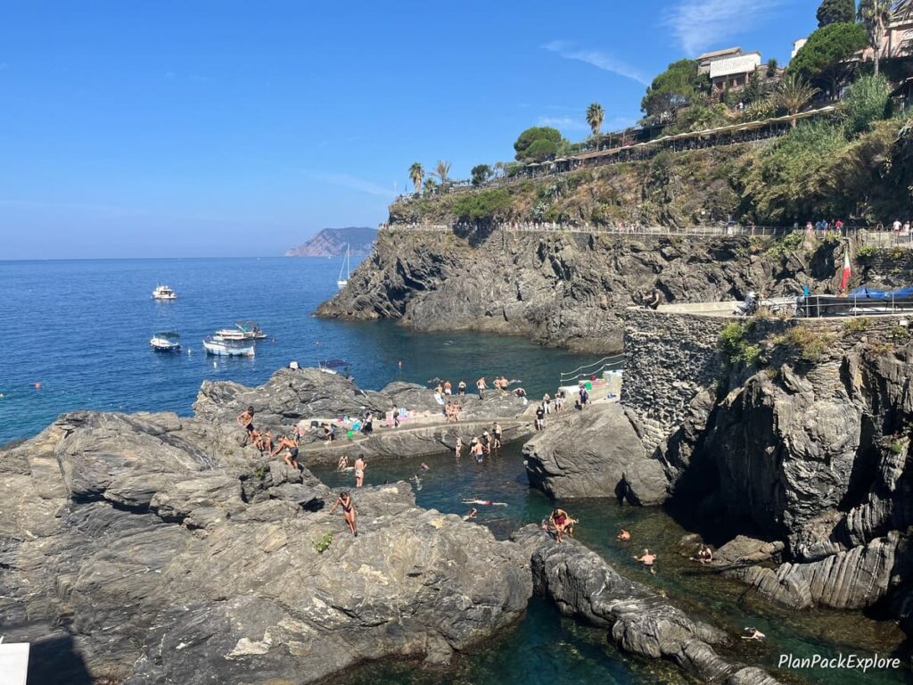 A large rock in the sea with people sunbathing on it and swimming, in Cinque Terre, Italy. Terraced vineyards on the cliff in the background.