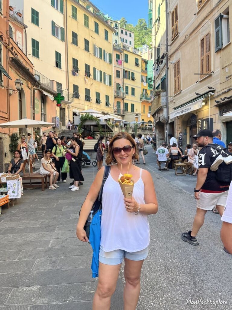 A person standing in the street of a village in Cinque Terre, eating seafood in a cone.