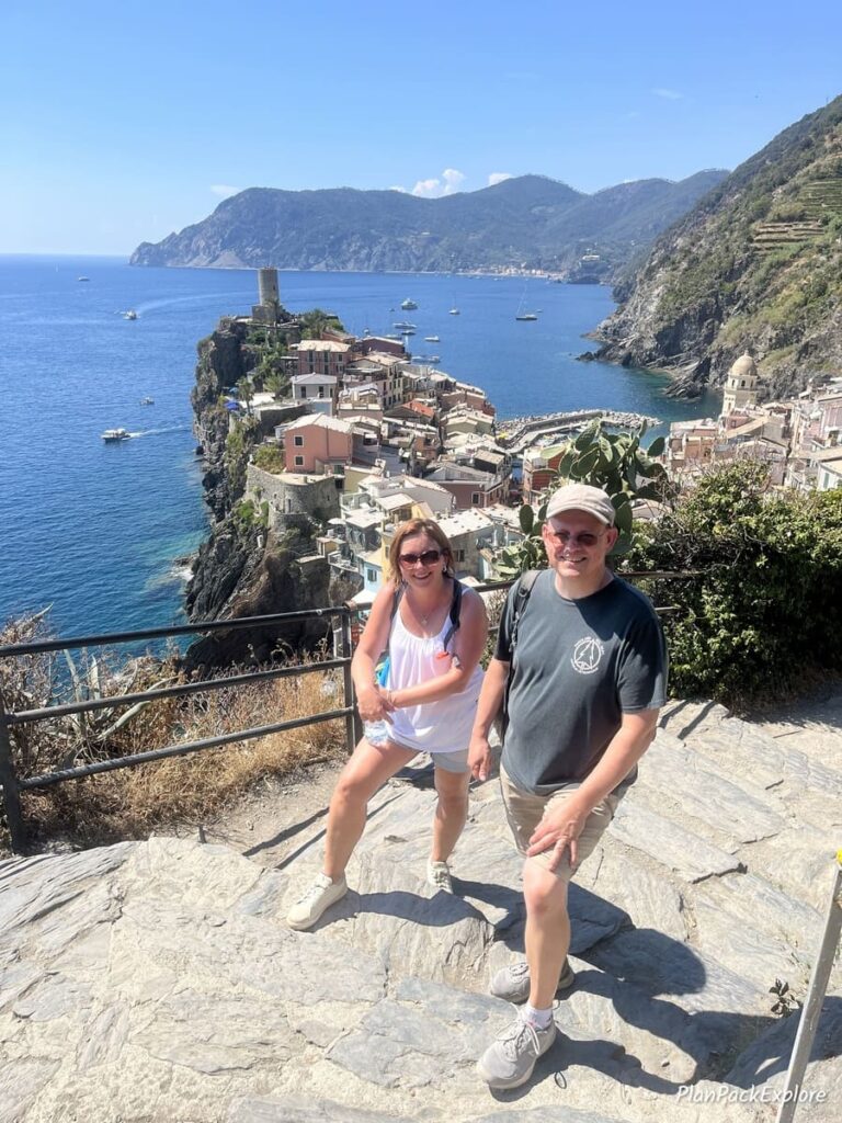 Two people posing for a picture on a trail in Cinque Terre, Italy, with the rooftops of the village of Vernazza and the sea in the background.