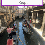 A gondola is navigated through a narrow canal in Venice, with colorful buildings lining the sides and tourists capturing photos alongside the water.