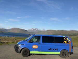 A blue campervan parked on a gravel lot with a view of a fjord and a mountain in Iceland.