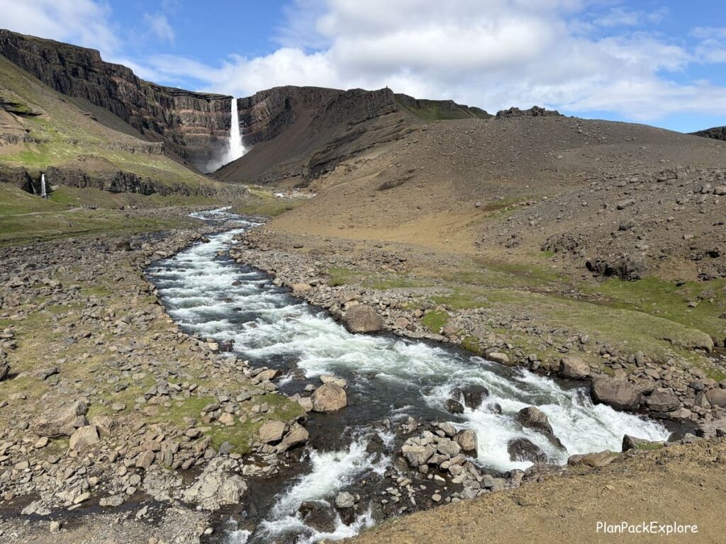 A tall waterfall in a dark basalt cove called Hengifoss, with a small river in front of it, in East Iceland.