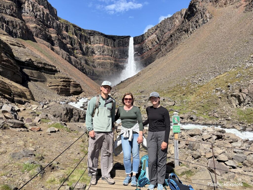 Three people standing in front of a tall waterfall nestled in a dark basalt cove with red horizontal striations called Hengifoss in East Iceland.