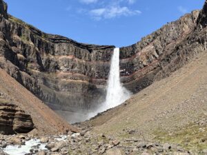 A tall waterfall in a dark basalt cove with red horizontal striations called Hengifoss in East Iceland.