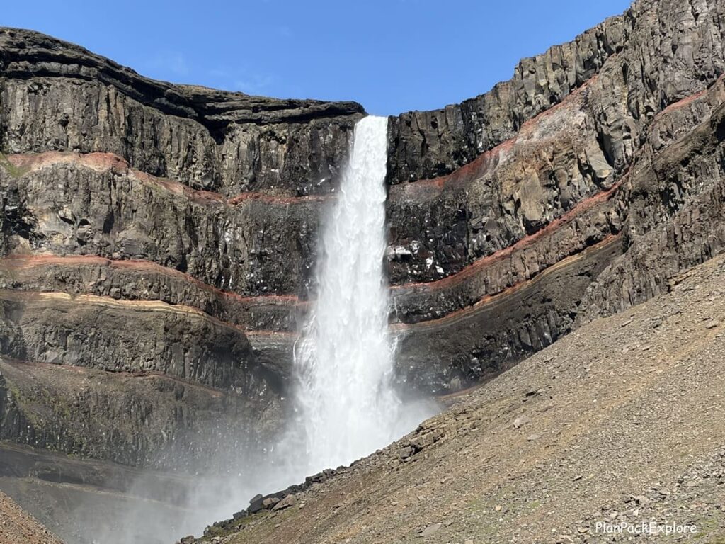 A close-up view of a tall waterfall in a dark basalt cove with red horizontal striations called Hengifoss in East Iceland.