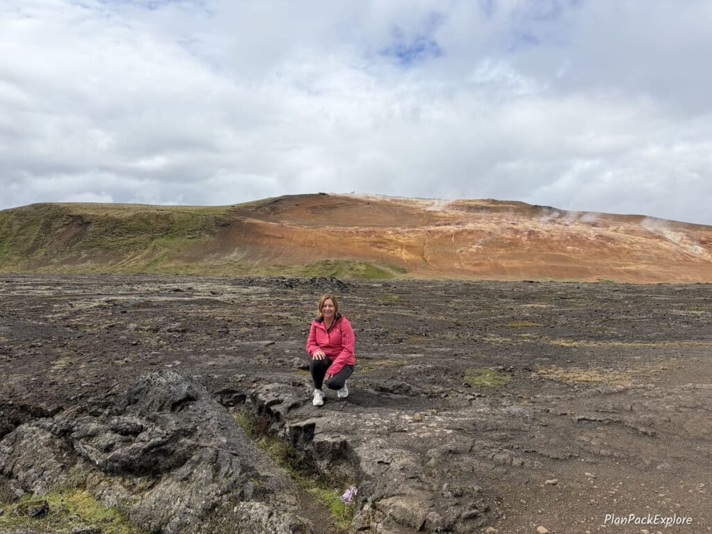 A person kneeling down for a photo on Leirhnjukur Lava Field, with a rhyolite mountain in the background.