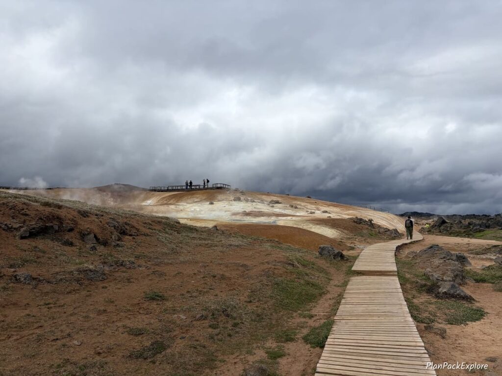 A wooden boardwalk next to a rhyolite hill with steam coming from the ground in the distance at Leirhnjukur in Iceland.