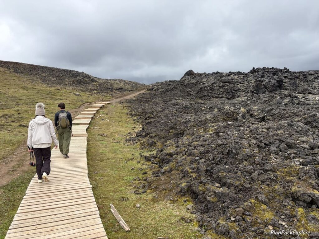 Two people walking on a wooden boardwalk next to a lava field at Leirhnjukur, Iceland.
