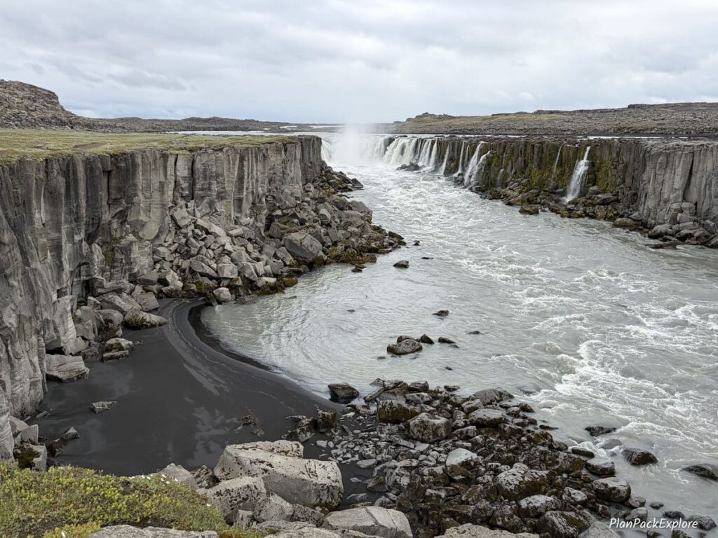 A small black sand beach area near a river and a waterfall in a canyon lined with basalt columns in Iceland.