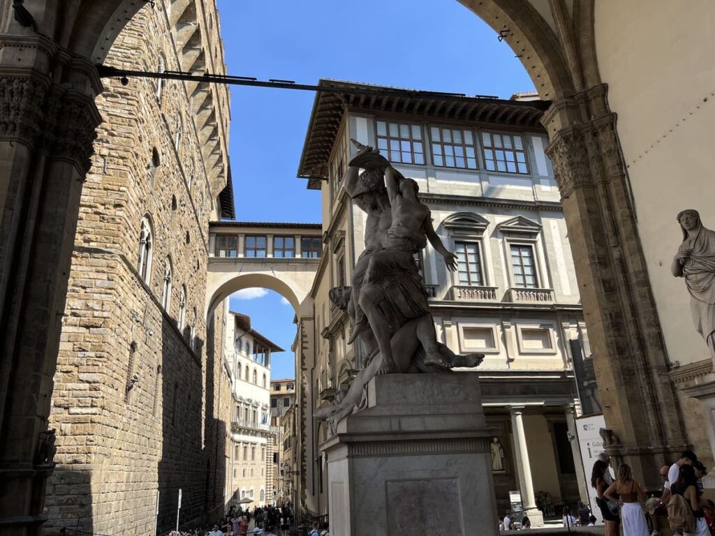 A view of a cream colored ornamental building with an arched covered passage of the Uffizi Galleries through the arch of Loggia dei Lanzi in Florence, Italy.