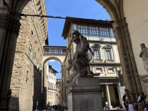 A view of a cream colored ornamental building with an arched covered passage of the Uffizi Galleries through the arch of Loggia dei Lanzi in Florence, Italy.